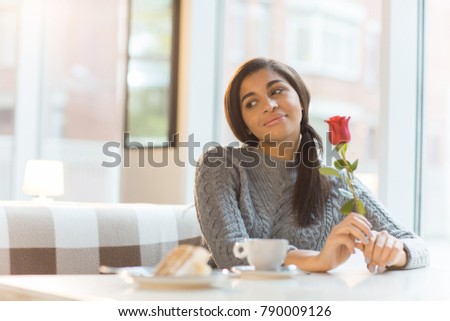 Happy young woman with fresh rose sitting by table in cafe and looking aside while thinking about her sweetheart