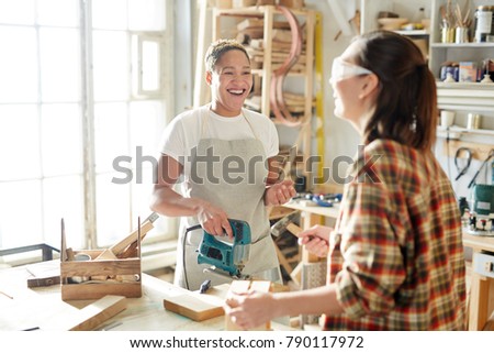 Laughing carpenter with powertool looking at her colleague during talk in workshop
