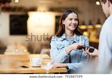Young smiling woman with card holding it over payment terminal while paying for her order in cafe