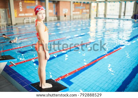 Cute schoolgirl in swimsuit, swim-cap and goggles looking at camera while standing on diving board