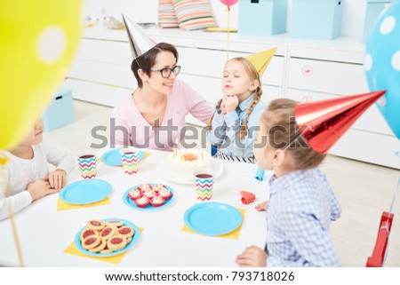 Cute girl making birthday wish while sitting by table and going to blow burning candles on cake