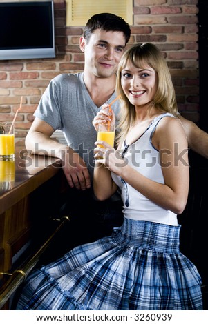 Smiling cute blond woman and handsome brunette man behind her turned back while drinking orange juice in the bar