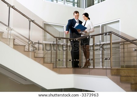 Young cute Hispanic businesswoman showing documents to her colleague on stairs in the office building
