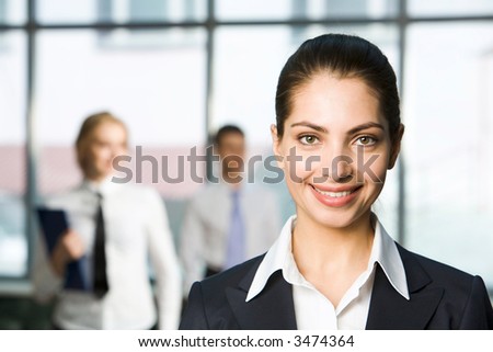 Portrait of pretty smiling brunette businesswoman looking at camera in the light office with large windows and two businesspeople on the background