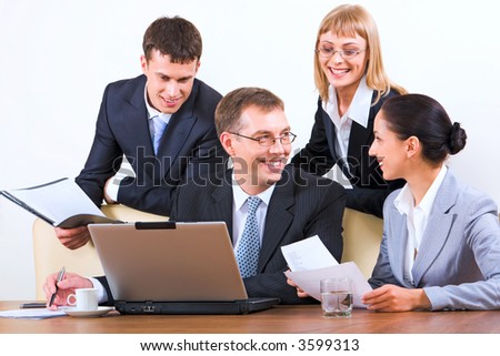 Group of four young businesspeople discussing different questions holding documents gathered together around the table with the laptop, drinks and papers on it