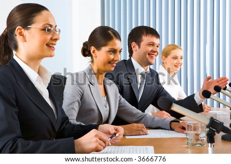 Four successful young people sitting in a line in front of monitors speaking on microphones sitting at the table in black comfortable chairs