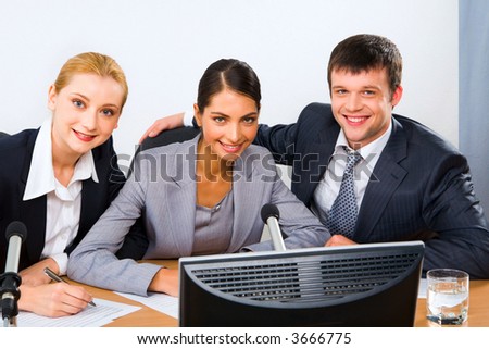 Three successful smiling colleagues sitting at the table with monitor, microphone and documents on it looking at the camera