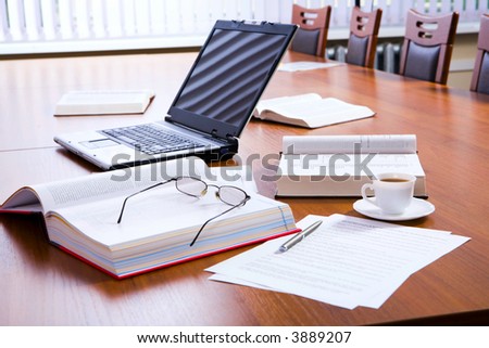 Opened thick books and laptop, documents, eyeglasses, pen and cup of coffee on large wooden table with row of chairs
