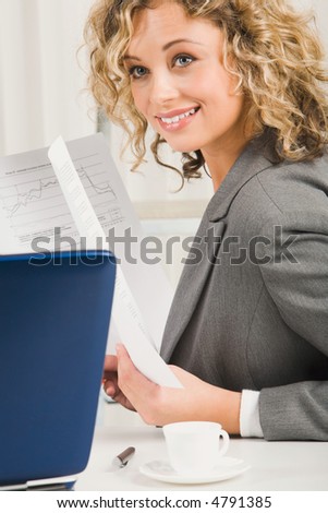 Portrait of curly cute business woman in gray jacket holding documents in her hands sitting at the table with cup, pen and opened paper case on it