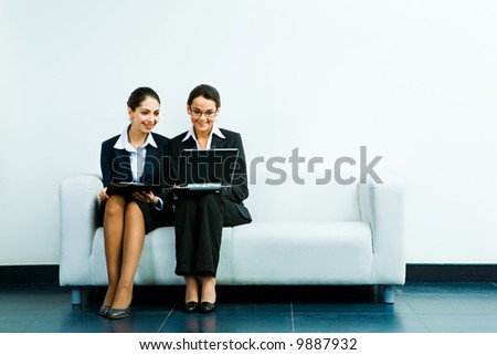 Portrait of business ladies sitting next to one another on the sofa and looking at the laptop screen with smiles