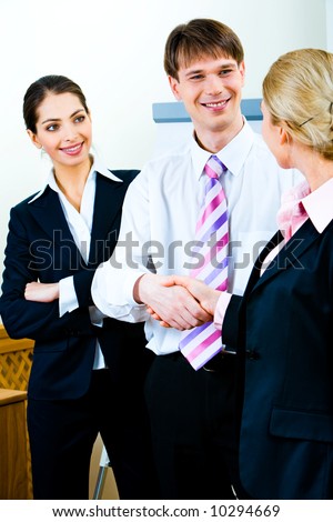 Portrait of smiling businessman shaking his co-worker’s hand at meeting while confident business lady in suit looking at them with smile