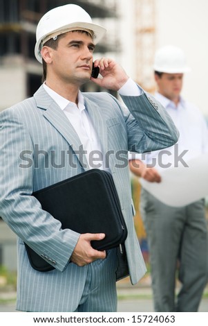 Portrait of busy engineer calling on his mobile phone at building site on the background of another worker