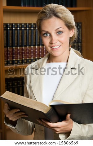 Portrait of elegant woman holding open book and looking at camera on background of shelves with tomes