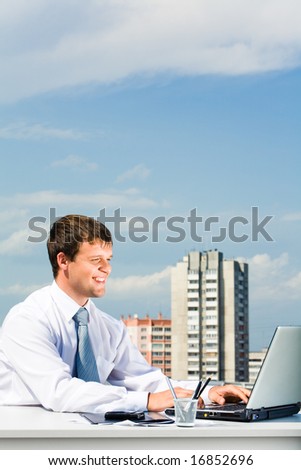 Portrait of smiling manager typing a letter on the laptop on the background of building with sky