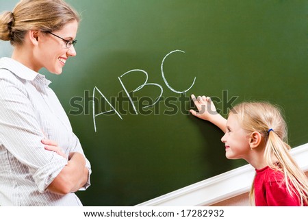Image of smart girl pointing at letter on blackboard and looking at her teacher with smile
