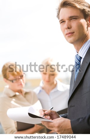 Portrait of serious boss holding pen over paper with working ladies at background