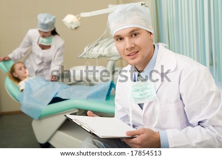 Image of handsome doctor with paper in hands looking at camera on background of assistant and little girl
