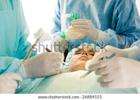 Photo of young female patient with closed eyes before operation with hands of surgeons over her holding surgical instruments and oxygen respirator