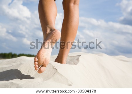 Close-up of slim legs of female walking down sandy beach with cloudy sky at background