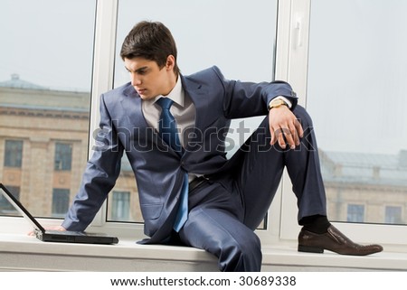 Photo of smart businessman sitting on window-sill with laptop near by and looking at monitor