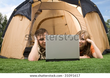Photo of modern couple lying on green grass and looking at laptop monitor in tent