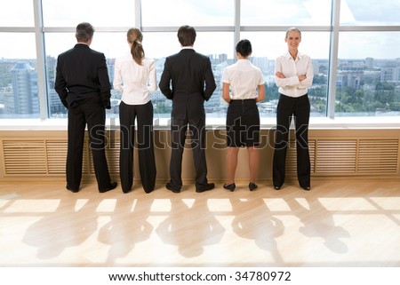Rear view of business group standing in row and looking through window with their leader smiling at camera