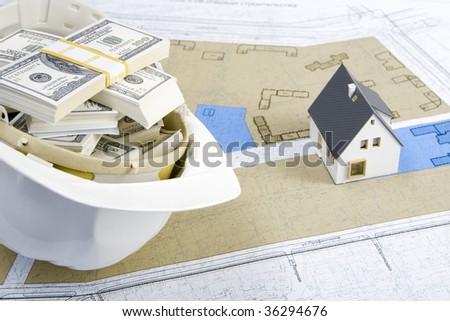 Close-up of toy house model on blueprints with helmet full of dollars near by