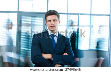 Portrait of confident man looking at camera in office with busy people passing by behind