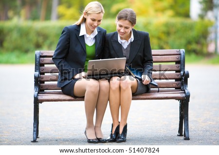 A portrait of two formally dressed women sitting on a bench with laptop