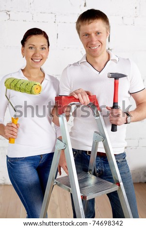 Industrious couple with paint roll and hammer looking at camera in new house
