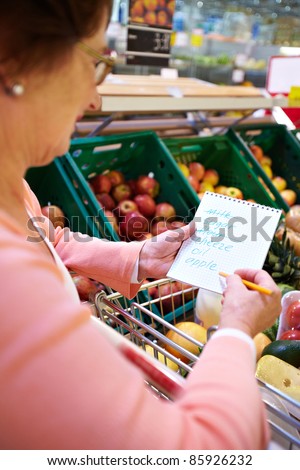 Image of senior woman looking at product list with goods in cart near by