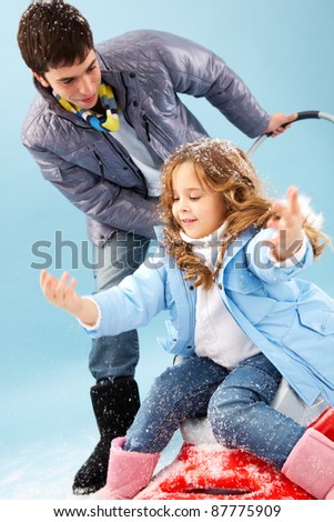 Happy girl sitting on sledge and catching snowflakes with her father near by