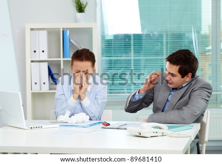 Image of businesswoman sneezing while her partner looking at her unsurely in office