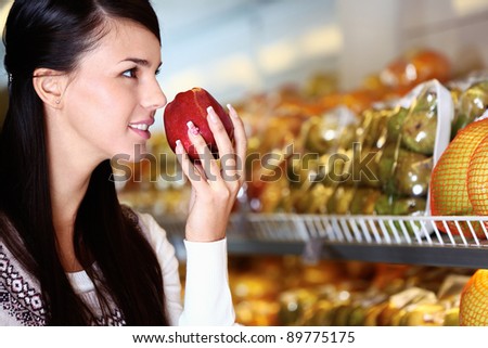 Image of young woman with fresh apple in hand smelling it in supermarket