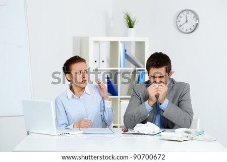 Image of sick businessman sneezing while anxious female looking at him in office