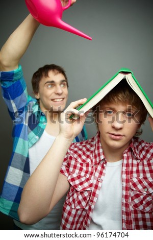 Cheerful guy pouring water from a pot while his friend using book as shelter, fool's day series
