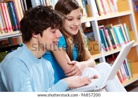 Guy using laptop at library while his girlfriend looking at screen with a smile
