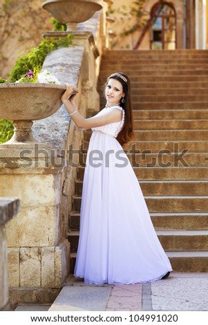 beautiful slim bride in luxury dress in park on sunset near blossom flowers in wedding day. young woman in Greek goddess style with diamond tiara and jewelery outdoor. romantic girl with glossy hair.
