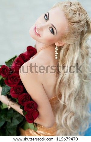 Beautiful blonde woman with red roses and long curly blond hair . Rich alluring girl in love dreaming and holding bouquet. lovely young lady with fresh flowers outdoor