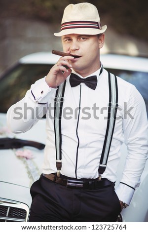 wedding - stylish groom posing outdoor with cigar and gangster hat. happy handsome man in suspenders and white shirt smoking and waiting for his bride - carnival costume wedding. smart italian mobster