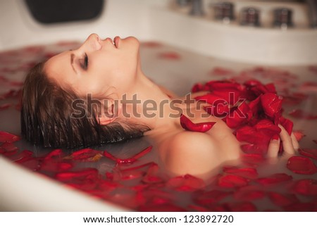 Attractive girl in bath with milk and rose petals. Spa treatments for skin rejuvenation. Alluring woman with bright makeup in Spa salon. Pretty sexy brunet relaxing in jacuzzi with red flower petals