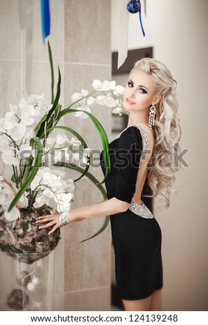 Beautiful slim woman with orchids in black dress in diamond jewelry with hairstyle and bright makeup. Sexy blond girl in fashion dress with flowers at hotel hall. Alluring model posing. tonality.