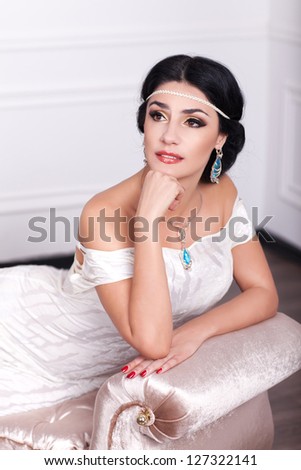 Woman in white dress resting in luxury inerrior - bride with bright makeup and fashion greek hairstyle in wedding dress and jewelry waiting for groom at home. stylish rich girl in vintage dress posing