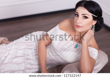Woman in white dress - bride with bright makeup and fashion greek hairstyle in wedding dress and jewelry waiting for groom at home. stylish girl in vintage dress have final preparation for wedding