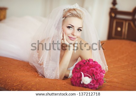 Gorgeous bride blonde with wedding bouquet makeup and hairstyle in bridal dress diamond jewelry at home waiting for groom. Jewelry and beauty. Rich happy girl  have final preparation for wedding