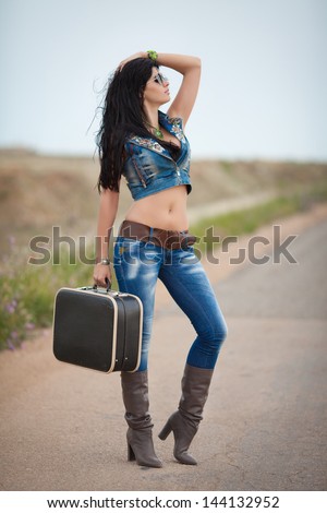 Attractive hiking woman hitchhiking along a road sexy Pretty brunette cowboy girl with travel bag. Happy smiling woman with suitcase at country road. Hiker portrait during hike trek.