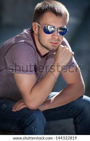 Handsome trendy man in fashion sunglasses in casual wear posing on city street in summer. Stylish smiling male model. Happy boy in fashion jeans waiting for girlfriend. thoughtful man in glasses
