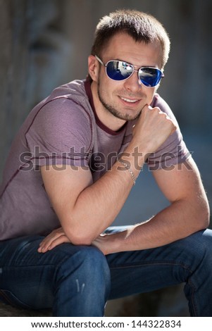 Handsome trendy man in fashion sunglasses in casual wear posing on city street in summer. Stylish smiling male model. Happy boy in fashion jeans waiting for girlfriend. thoughtful man in glasses