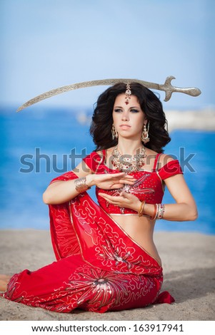 Beautiful Indian woman dancing with golden sword outdoors. Indian dancer girl in traditional red dress in India. Portrait of young Indian woman dancing at sea beach.