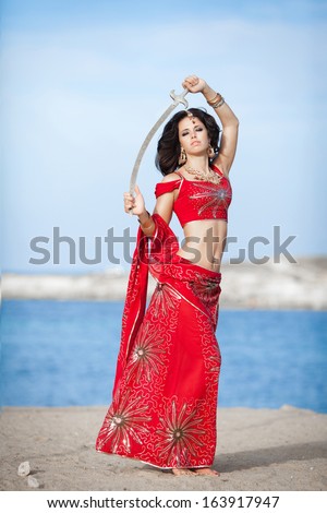 Beautiful Indian woman dancing with golden sword outdoors. Indian dancer girl in traditional red dress in India. Portrait of young Indian woman dancing at sea beach.